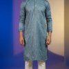 Men's dusty blue Lucknowi Chikankari kurta with intricate embroidery and mirror work, paired with white trousers and tan sandals (1)