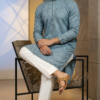 Men's dusty blue Lucknowi Chikankari kurta with intricate embroidery and mirror work, paired with white trousers and tan sandals (1)