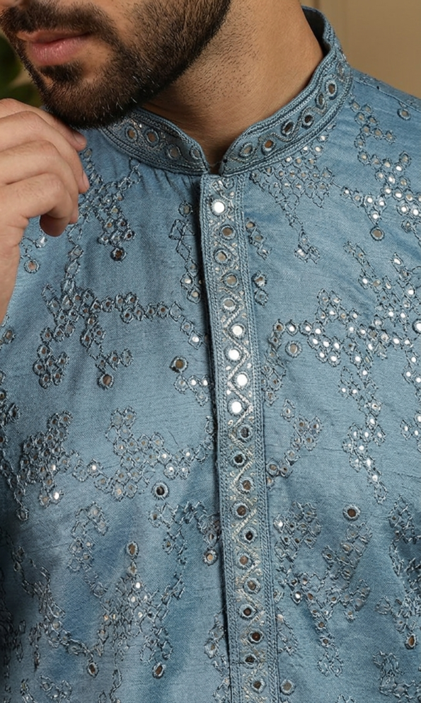 Men's dusty blue Lucknowi Chikankari kurta with intricate embroidery and mirror work, paired with white trousers and tan sandals (1)
